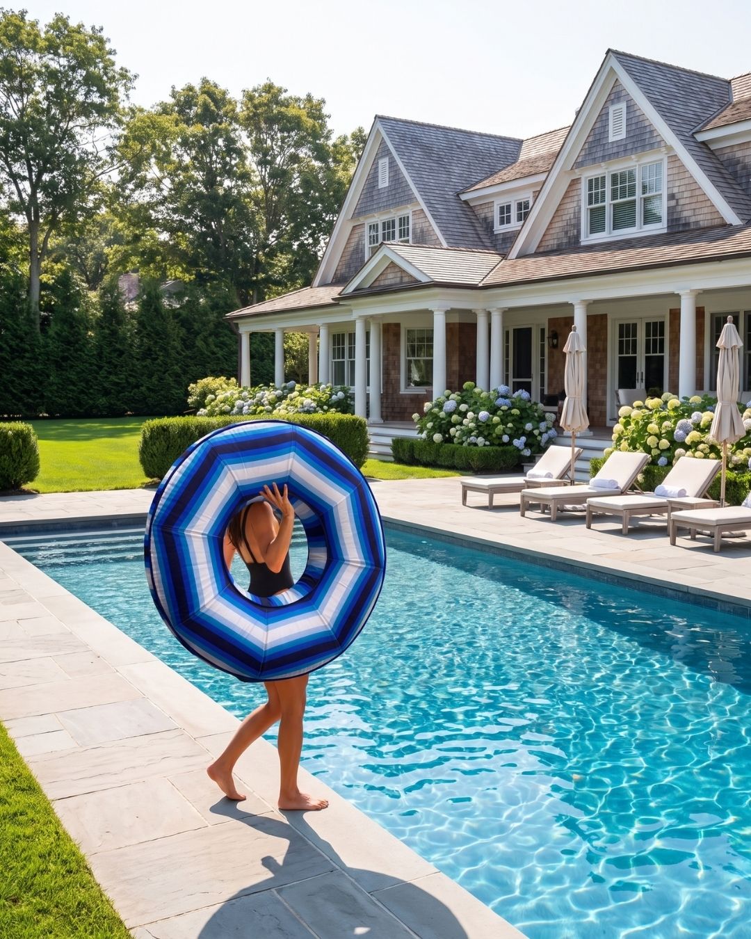 A woman in a black swimsuit walks beside a swimming pool holding a blue and white striped inflatable pool float ring in front of a large house with a porch and manicured lawn.