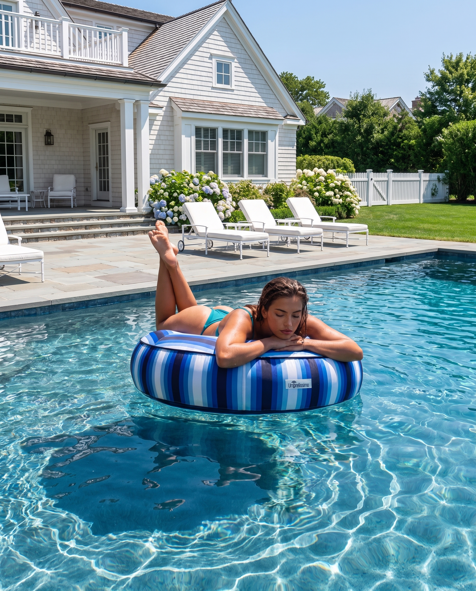 A woman relaxes on a blue and white striped luxury umbrellissimo inflatable pool float in a swimming pool, with a large white house, lounge chairs, and green lawn in the background.