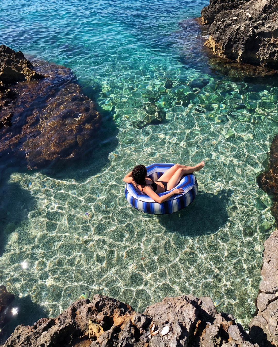 A woman relaxes in a blue and white striped inflatable pool float on clear turquoise water surrounded by dark rocky cliffs.