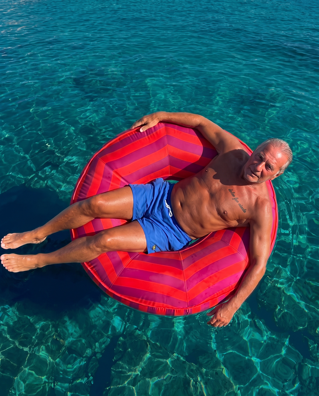 A man lounging on a red and fuchsia striped upholstered inflatable pool float in clear turquoise ocean pool water.