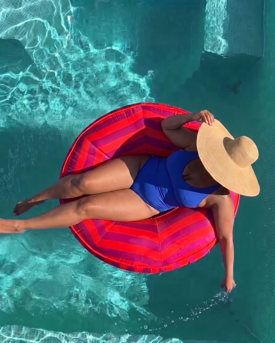 A woman wearing a blue swimsuit and a large straw sun hat lounges on a red and fuchsia striped inflatable pool float in a swimming pool.